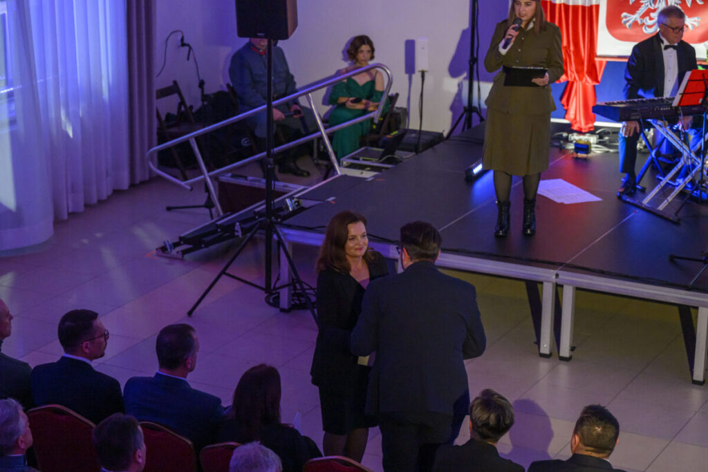 Na zdjęciu widać scenę z kobietą przemawiającą do mikrofonu. Publiczność siedzi na krzesłach, wokół ciemniejsze oświetlenie. W tle czerwone zasłony i godło.