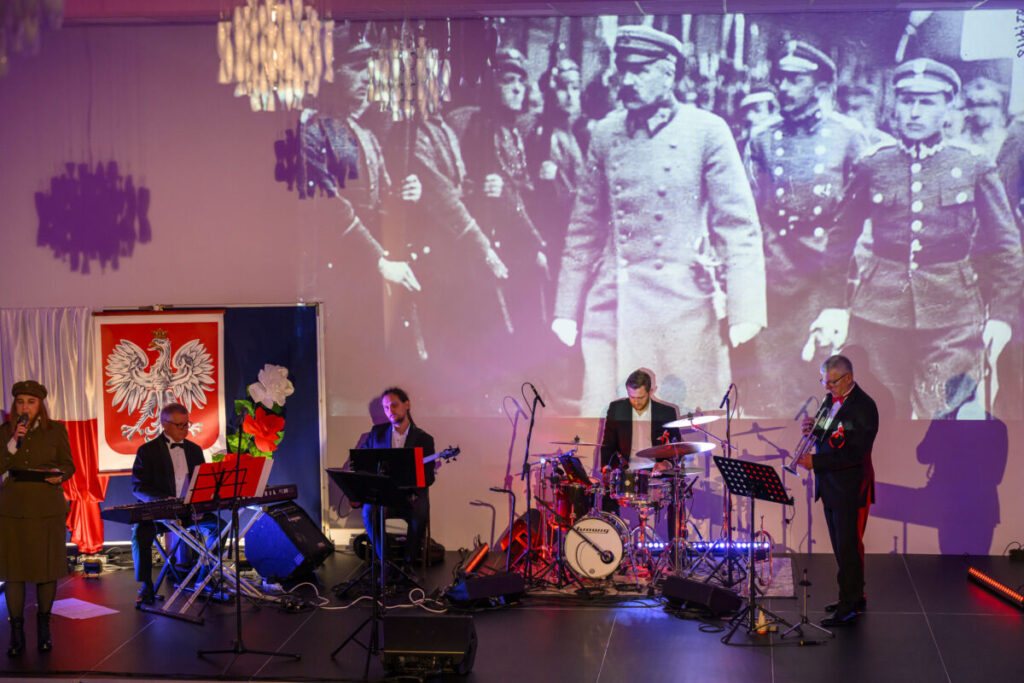 Zespół muzyczny gra na scenie z wyświetlanym na ścianie historycznym zdjęciem żołnierzy. W tle biało-czerwona flaga z godłem Polski. Oświetlenie sceniczne jest w odcieniach fioletu i czerwieni.
