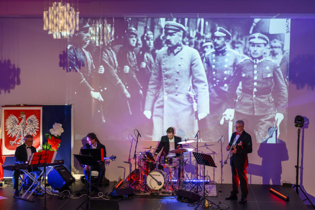 Grupa muzyków gra na scenie z niebieskim oświetleniem. W tle duża, czarno-biała projekcja historycznych żołnierzy. Obok widać polskie godło na czerwonym tle.