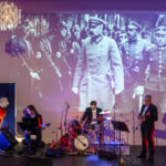 Grupa muzyków gra na scenie z niebieskim oświetleniem. W tle duża, czarno-biała projekcja historycznych żołnierzy. Obok widać polskie godło na czerwonym tle.