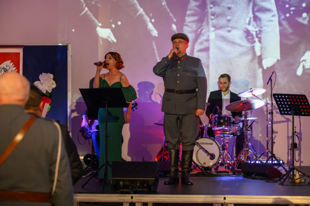 Osoby śpiewające na scenie w eleganckich strojach, tło to biało-czarny obraz historyczny, zespół muzyczny z perkusją.