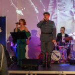 Osoby śpiewające na scenie w eleganckich strojach, tło to biało-czarny obraz historyczny, zespół muzyczny z perkusją.