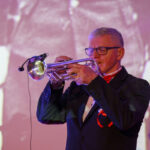 Mężczyzna w garniturze gra na trąbce. Tło jest rozmyte, w odcieniach fioletu i różu. Muzyk nosi okulary i czerwony krawat.