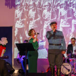 Koncert przed historycznym zdjęciem żołnierzy, śpiewają kobieta w zielonej sukni i mężczyzna w mundurze, w tle perkusista.