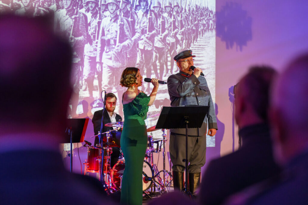 Na scenie kobieta i mężczyzna śpiewają, z tyłu perkusista. Kobieta w zielonej sukni, mężczyzna w mundurze. Tło to czarno-białe zdjęcie żołnierzy.