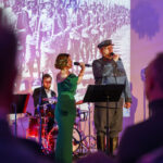 Na scenie kobieta i mężczyzna śpiewają, z tyłu perkusista. Kobieta w zielonej sukni, mężczyzna w mundurze. Tło to czarno-białe zdjęcie żołnierzy.