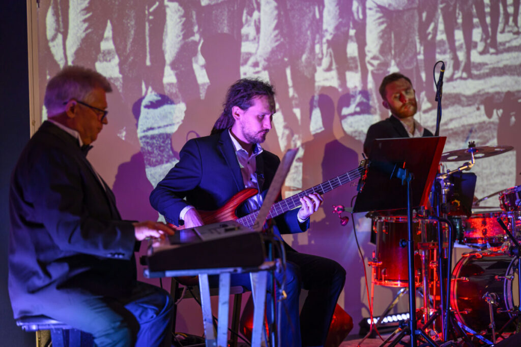 Trzech muzyków gra: jeden na klawiszach, drugi na gitarze basowej, trzeci na perkusji. Tło z projekcją w sepii. Oświetlenie purpurowe i czerwone.