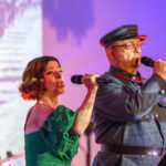 Dwoje ludzi śpiewa na scenie; kobieta w zielonej sukni i mężczyzna w mundurze. W tle różowe oświetlenie i historyczne zdjęcie.