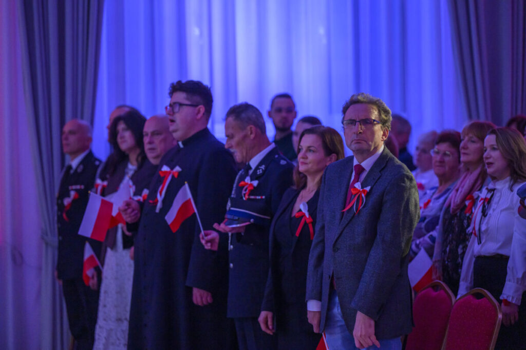 Grupa ludzi w sali z niebieskim oświetleniem, trzymających polskie flagi. Mają przypięte czerwono-białe kokardy. Tło z zasłonami.