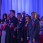 Grupa ludzi w sali z niebieskim oświetleniem, trzymających polskie flagi. Mają przypięte czerwono-białe kokardy. Tło z zasłonami.