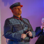 Na zdjęciu mężczyzna w historycznym wojskowym mundurze, z mikrofonem w dłoni. Tło jest rozmyte, oświetlone na kolorowo. Obok widoczny jest drugi mężczyzna grający na trąbce.