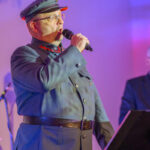 Mężczyzna w granatowym mundurze z czapką śpiewa do mikrofonu. Tło w odcieniach fioletu. Obok widoczny statyw.