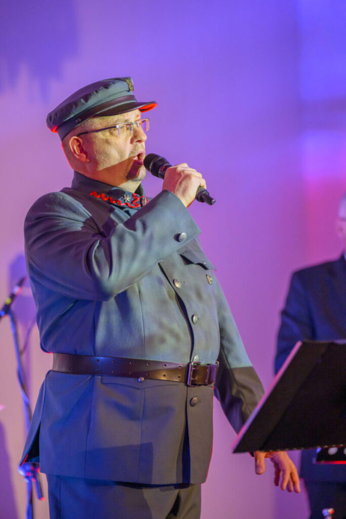 Mężczyzna w granatowym mundurze z czapką śpiewa do mikrofonu. Tło w odcieniach fioletu. Obok widoczny statyw.