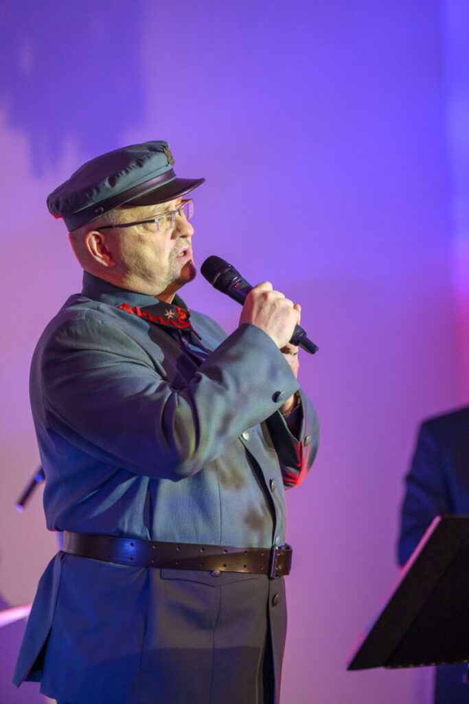 Osoba w mundurze śpiewa do mikrofonu. Scena oświetlona na fioletowo i różowo. Tło jednolite, zaciemnione.