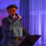 Mężczyzna w mundurze wojskowym mówi do mikrofonu. Tło oświetlone jest na niebiesko i fioletowo, z lekkimi pionowymi pasmami. Na pierwszym planie stojak na nuty.