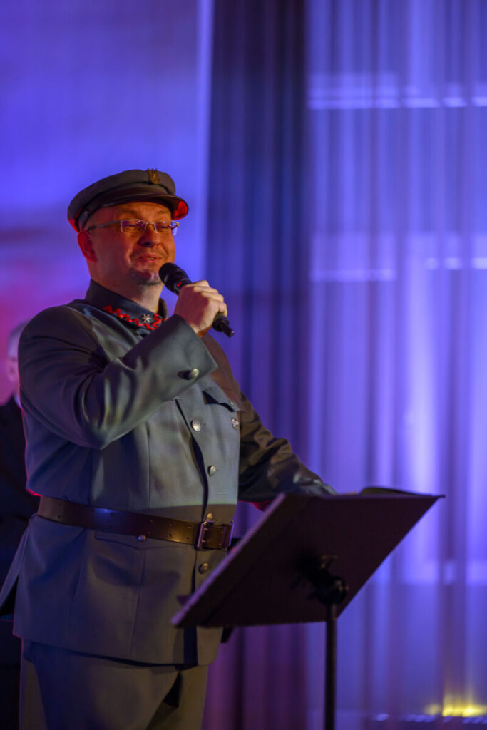 Mężczyzna w mundurze wojskowym mówi do mikrofonu. Tło oświetlone jest na niebiesko i fioletowo, z lekkimi pionowymi pasmami. Na pierwszym planie stojak na nuty.