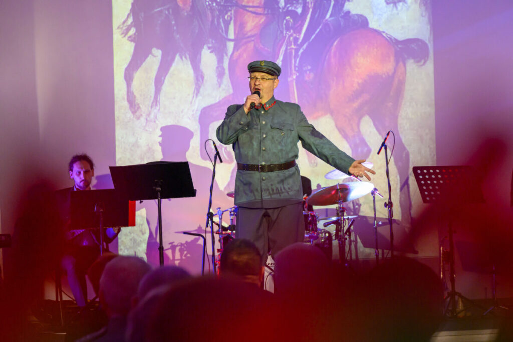 Mężczyzna w mundurze śpiewa na scenie; za nim obraz z koniem. Tło w pastelowych kolorach, sceniczne oświetlenie.