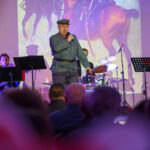 Na scenie występuje osoba w mundurze, gra muzyka. Tło to obraz konia, a kolorystyka dominuje w ciemnych barwach. Widać kilka instrumentów muzycznych i publiczność.