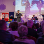 Na scenie odbywa się koncert; artysta śpiewa, a zespół gra. W tle biało-czerwona flaga i obraz z żołnierzem i koniem. Publiczność siedzi na widowni.