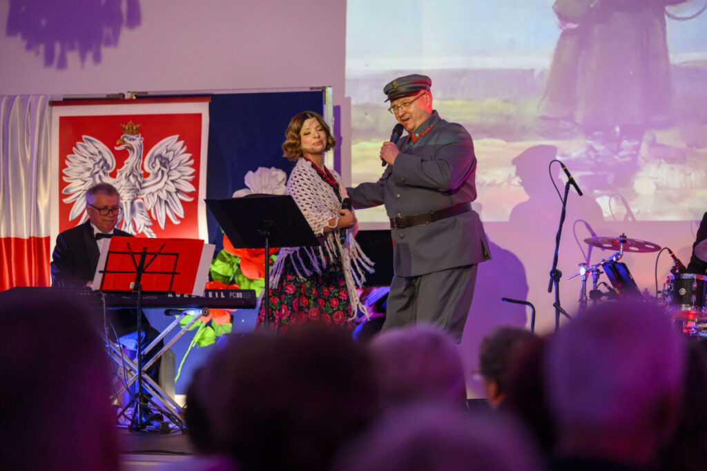 Dwoje ludzi na scenie, mężczyzna w mundurze śpiewa do mikrofonu. W tle biało-czerwona flaga i herb Polski. Publiczność w cieniu, kolorowe oświetlenie.