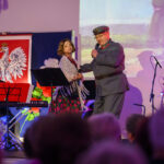 Dwoje ludzi na scenie, mężczyzna w mundurze śpiewa do mikrofonu. W tle biało-czerwona flaga i herb Polski. Publiczność w cieniu, kolorowe oświetlenie.