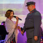 Na scenie kobieta z mikrofonem i mężczyzna w mundurze, przy kolorowym tle. Kobieta nosi białą chustę. Mężczyzna ma szary mundur i czapkę.