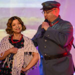 Na zdjęciu są dwie osoby na scenie, trzymają mikrofony. Kobieta ma jasny szal i korale, mężczyzna jest w mundurze. Tło jest kolorowe, z elementami przyrody.