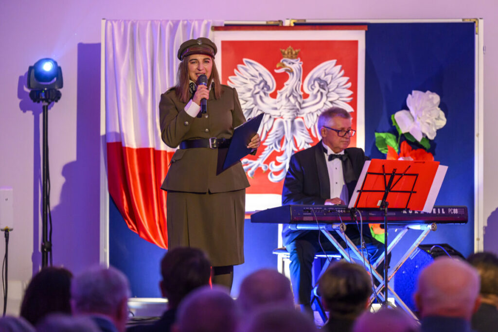 Na scenie kobieta w mundurze mówi do mikrofonu, obok niej mężczyzna gra na keyboardzie. W tle znajduje się flaga Polski i godło. Dominuje czerwony, biały i niebieski kolor.