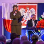 Na scenie kobieta w mundurze mówi do mikrofonu, obok niej mężczyzna gra na keyboardzie. W tle znajduje się flaga Polski i godło. Dominuje czerwony, biały i niebieski kolor.