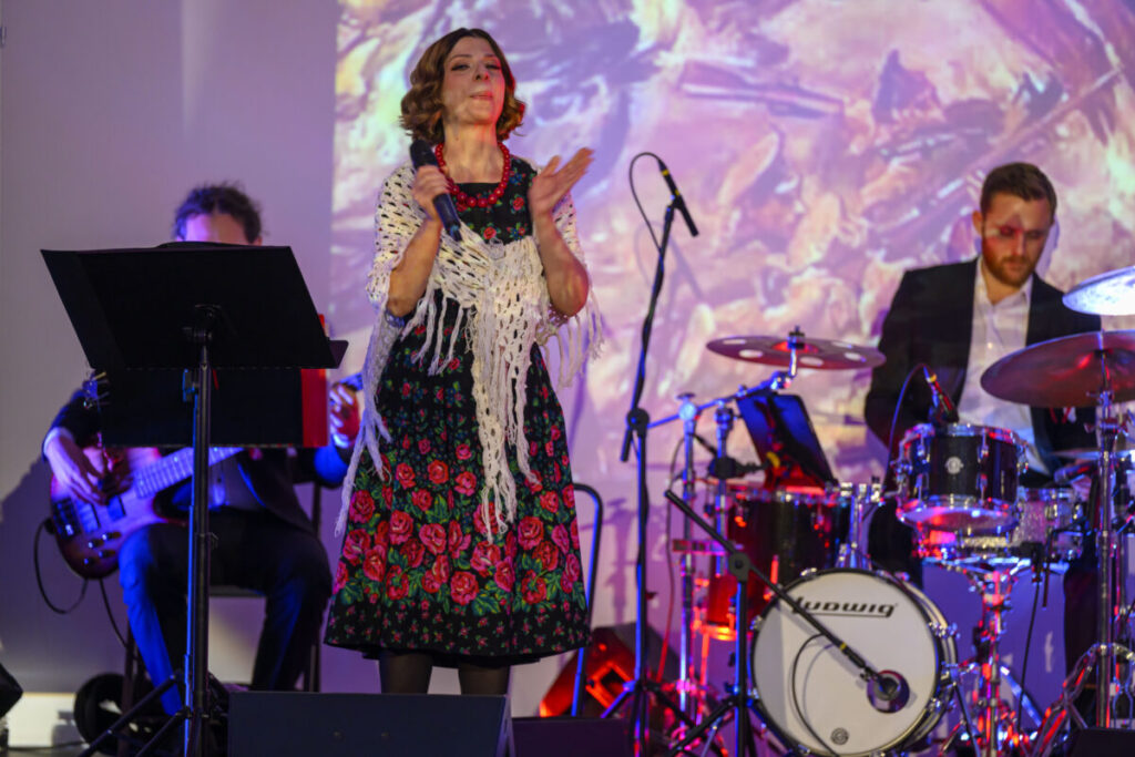 Na scenie występuje kobieta w kolorowej sukience, obok perkusista. Tło jasne, abstrakcyjne wzory. Instrumenty i mikrofon na przodzie.