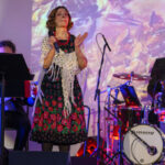 Na scenie występuje kobieta w kolorowej sukience, obok perkusista. Tło jasne, abstrakcyjne wzory. Instrumenty i mikrofon na przodzie.