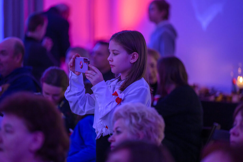 Dziewczynka w białej bluzce fotografuje telefonem. Jasne tło w odcieniach różu i fioletu. W tle niewyraźne sylwetki ludzi.
