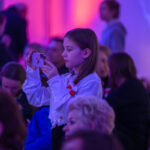 Dziewczynka w białej bluzce fotografuje telefonem. Jasne tło w odcieniach różu i fioletu. W tle niewyraźne sylwetki ludzi.