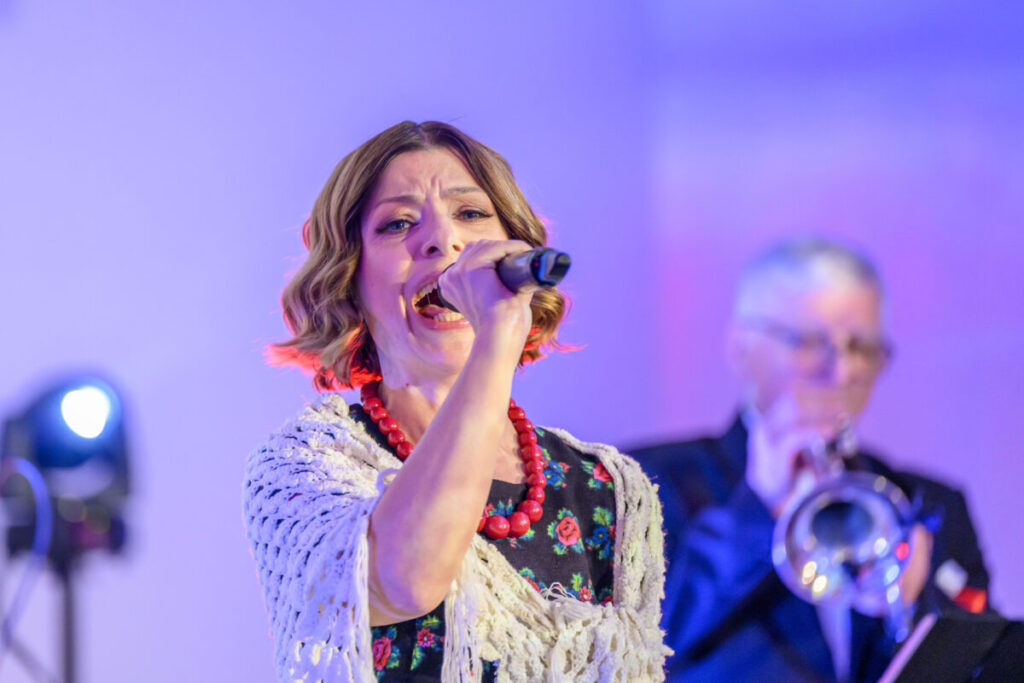 Na zdjęciu młoda kobieta śpiewa do mikrofonu, ma na sobie kolorową sukienkę i biały szal. Tło jest fioletowo-niebieskie z oświetleniem scenicznym. Obok niej w oddali widać mężczyznę grającego na trąbce.