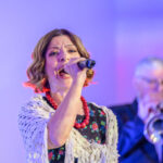 Na zdjęciu młoda kobieta śpiewa do mikrofonu, ma na sobie kolorową sukienkę i biały szal. Tło jest fioletowo-niebieskie z oświetleniem scenicznym. Obok niej w oddali widać mężczyznę grającego na trąbce.