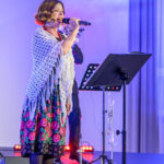 Kobieta śpiewa na scenie, ubrana w folkową sukienkę z kwiatami i białą chustę. Tło jest niebieskie, z muzykami w tle. Światła sceniczne emitują ciepłe barwy.