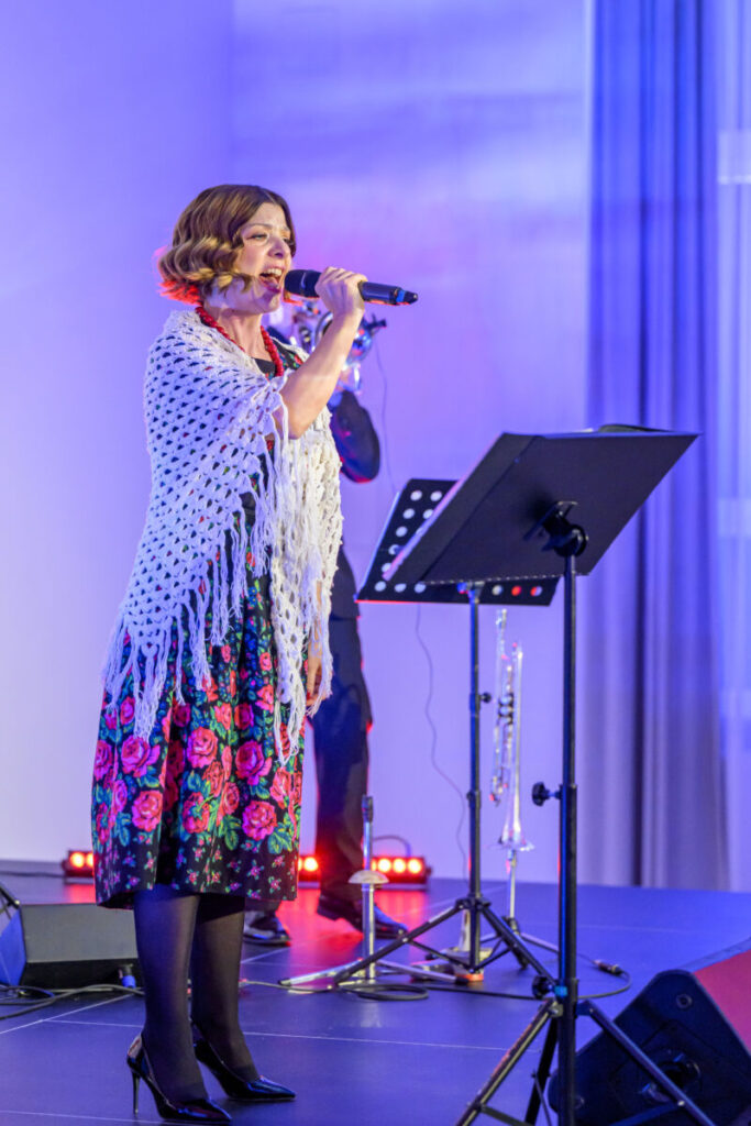 Kobieta śpiewa na scenie, ubrana w folkową sukienkę z kwiatami i białą chustę. Tło jest niebieskie, z muzykami w tle. Światła sceniczne emitują ciepłe barwy.