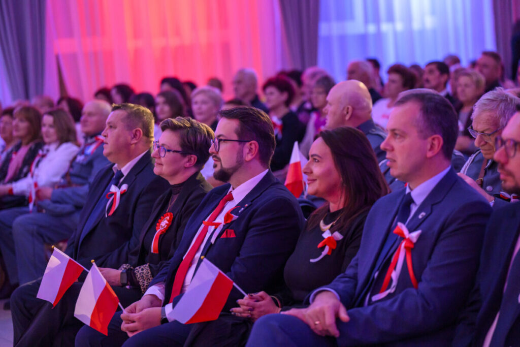 Grupa ludzi siedzi w rzędu podczas wydarzenia, trzymając polskie flagi. Dominują kolory biały, czerwony i niebieski. Tło jest rozmyte ze względu na oświetlenie.