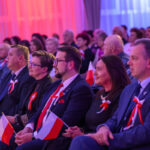 Grupa ludzi siedzi w rzędu podczas wydarzenia, trzymając polskie flagi. Dominują kolory biały, czerwony i niebieski. Tło jest rozmyte ze względu na oświetlenie.