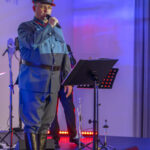 Mężczyzna w niebieskim mundurze śpiewa na scenie. Tło jest oświetlone na niebiesko i czerwono. Obok mikrofon, w tle instrumenty.