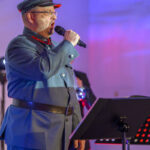 Mężczyzna w mundurze wojskowym śpiewa do mikrofonu. Tło jest niebiesko-czerwone, obok stojak na nuty.