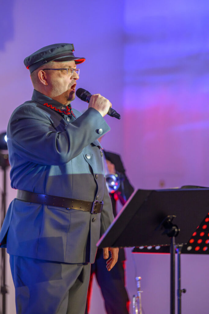 Mężczyzna w mundurze wojskowym śpiewa do mikrofonu. Tło jest niebiesko-czerwone, obok stojak na nuty.