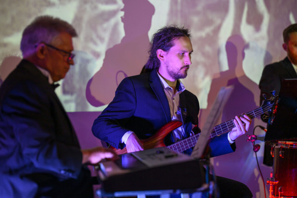 Na zdjęciu trzej mężczyźni grają w ciemnych garniturach na instrumentach w jasnym pokoju. Ściana w tle ma kolorowe światła. Klawisze, gitara i sprzęt muzyczny są widoczne.