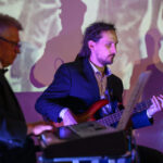 Na zdjęciu trzej mężczyźni grają w ciemnych garniturach na instrumentach w jasnym pokoju. Ściana w tle ma kolorowe światła. Klawisze, gitara i sprzęt muzyczny są widoczne.