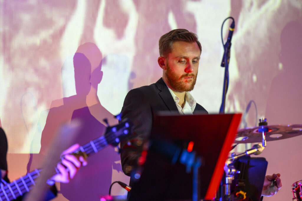 Muzyk gra na perkusji w kolorowym świetle. Ma krótkie włosy i brodę, nosi ciemny garnitur. Tło z abstrakcyjnymi wzorami.