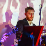 Muzyk gra na perkusji w kolorowym świetle. Ma krótkie włosy i brodę, nosi ciemny garnitur. Tło z abstrakcyjnymi wzorami.