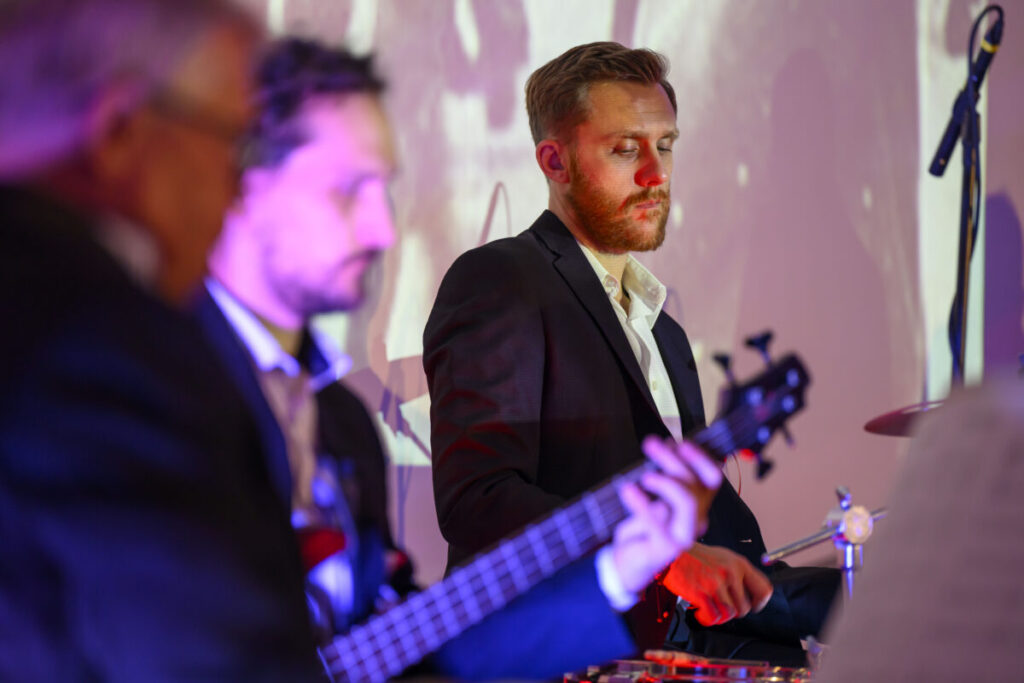 Na zdjęciu widać muzyków na scenie w eleganckich garniturach. Mężczyzna z brodą gra na perkusji, drugi na gitarze. Tło jest jasne, z fioletowym oświetleniem.