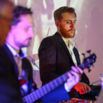 Na zdjęciu widać muzyków na scenie w eleganckich garniturach. Mężczyzna z brodą gra na perkusji, drugi na gitarze. Tło jest jasne, z fioletowym oświetleniem.