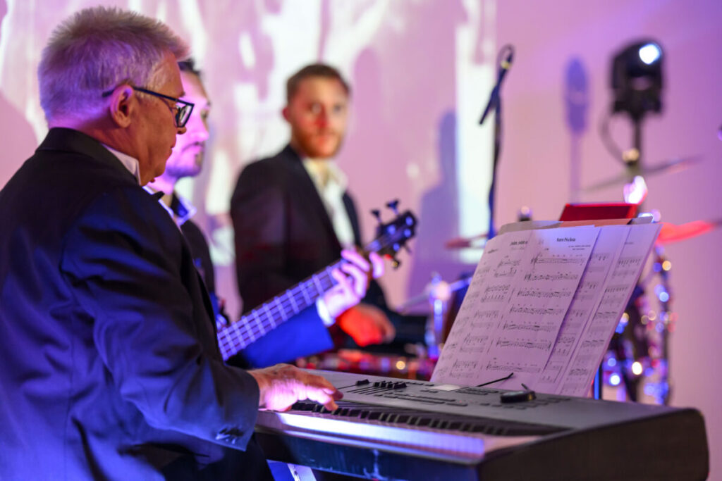 Trzej mężczyźni w garniturach grają na keyboardzie i basie. Scena podświetlona jest na fioletowo. W tle stoją instrumenty perkusyjne.
