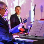 Trzej mężczyźni w garniturach grają na keyboardzie i basie. Scena podświetlona jest na fioletowo. W tle stoją instrumenty perkusyjne.